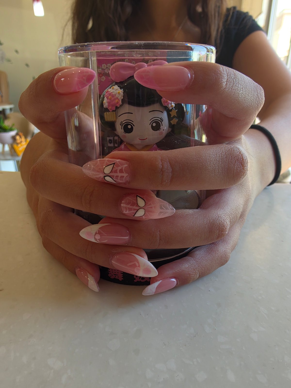 Two hands with long almond-shaped nails in soft pink ombré, hand-painted white spiderwebs and glossy white tips, holding a clear cup.